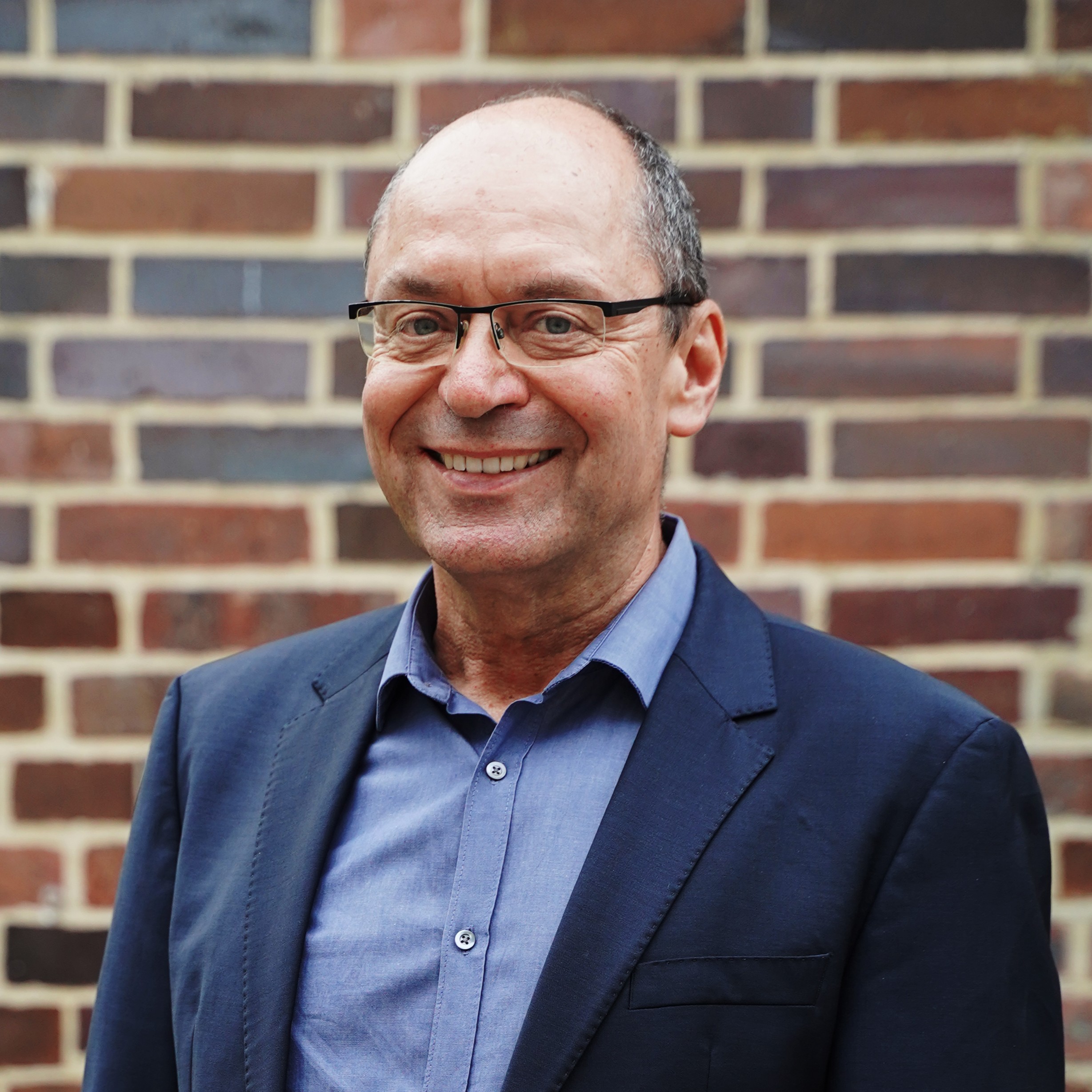 Porträtfoto von Marcel Keiffenheim: Ein lächelnder Mann mit grauem Haar, Halbglatze und Brille, der ein dunkelblaues Jackett und ein hellblaues Hemd trägt.