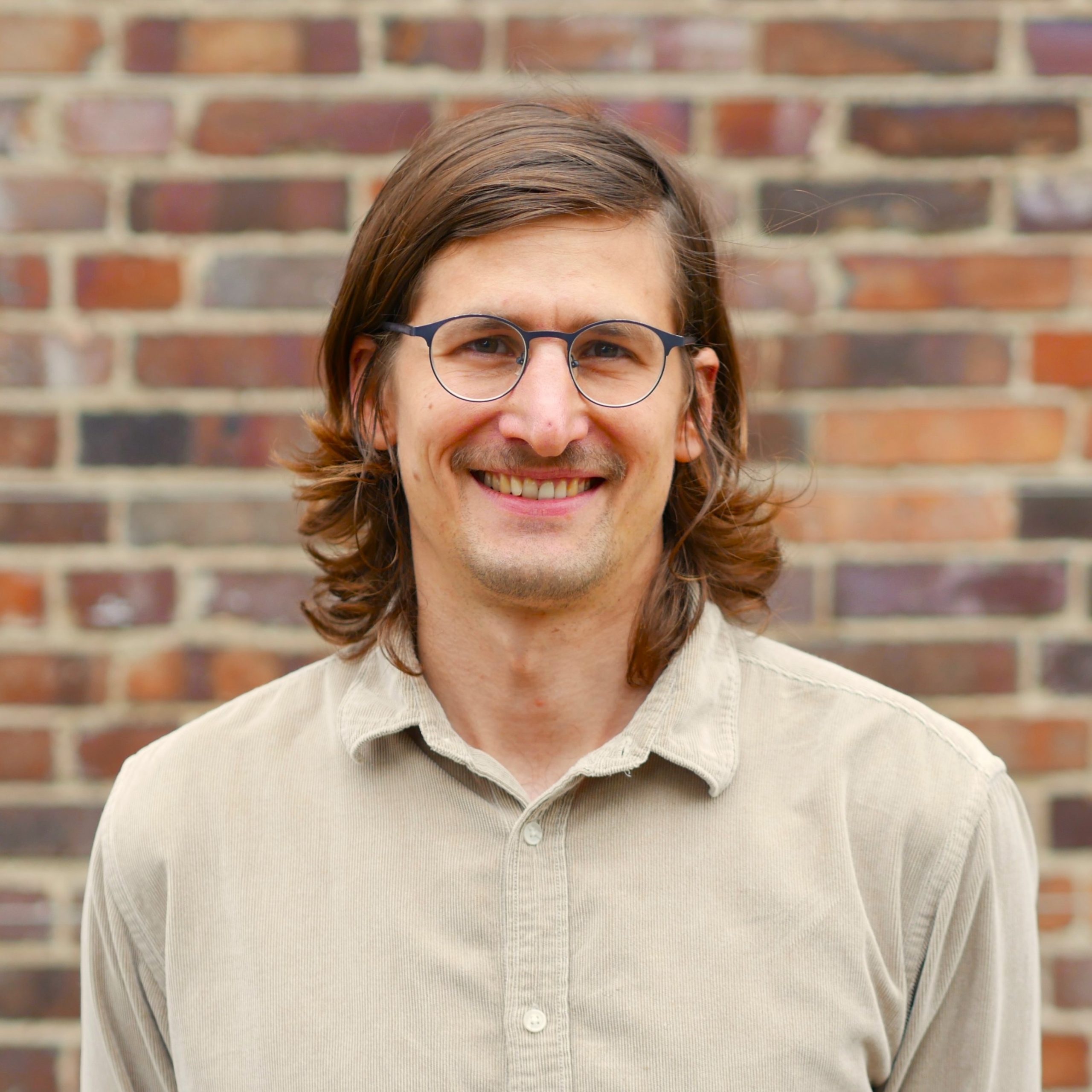 Portrait photo of Paul Mougeolle: a smiling young man with shoulder-length brown hair and glasses.