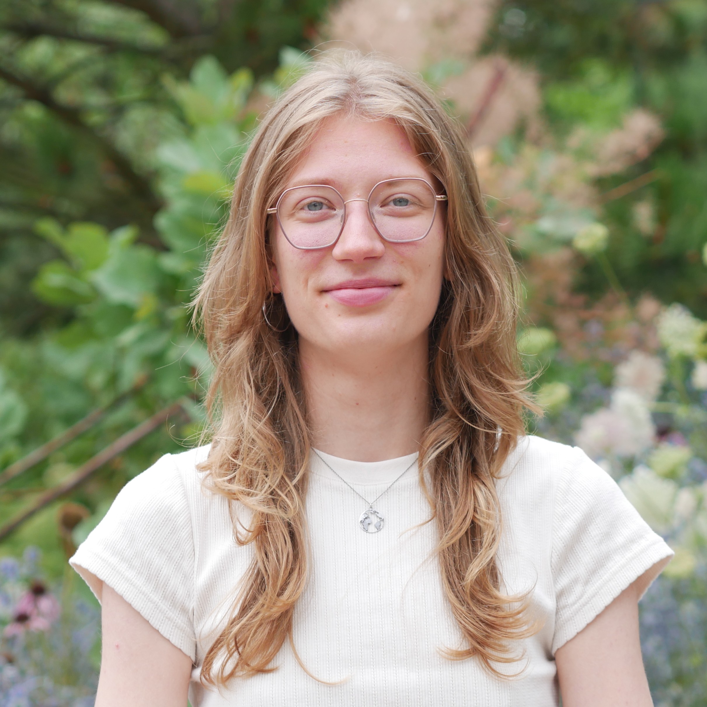 Portrait photo of Kora Stehr: A young blonde woman wearing glasses against the backdrop of a garden landscape.