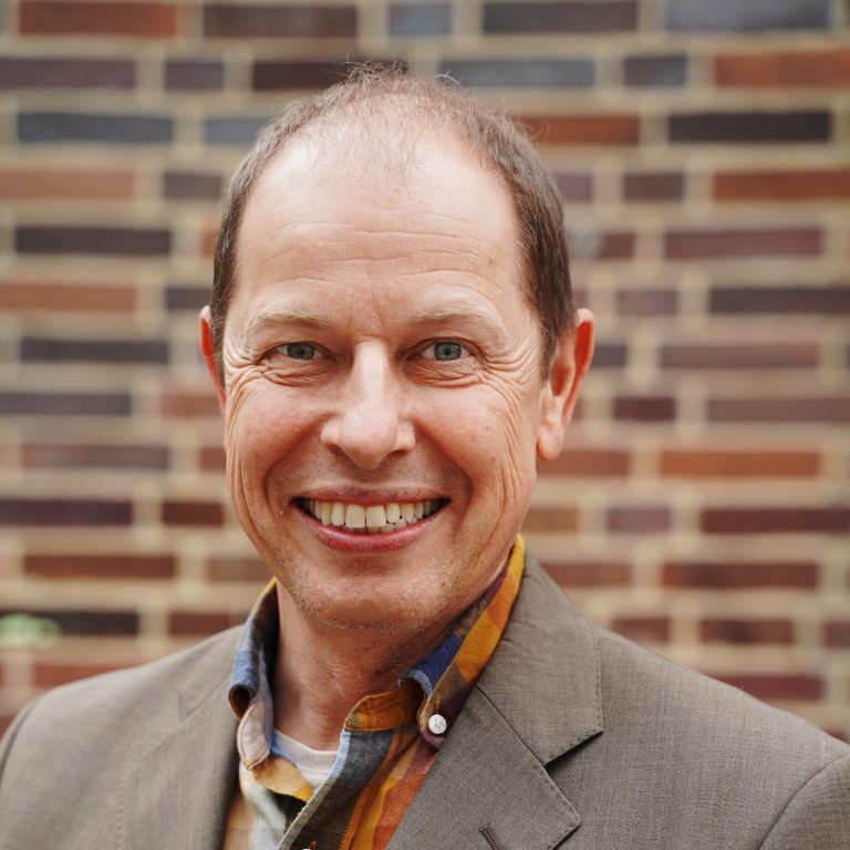 Porträtfoto von Tobias Ott: Ein grinsender Mann mit braunem Haar und Halbglatze, der ein buntes Hemd und ein graubraunes Jackett trägt.