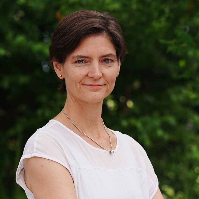 Porträtaufnahme von Henrike Lindemann: Eine lächelnde Frau mit kurzem braunen Haar und graublauen Augen, die eine Silberkette und ein weißes T-shirt trägt.