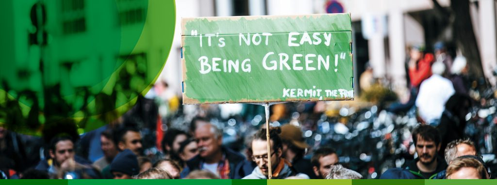 Bei einer Klimademonstration halten Menschen ein grünes Pappschild mit der Aufschrift: "It's not easy being green! - Kermit, the Frog"