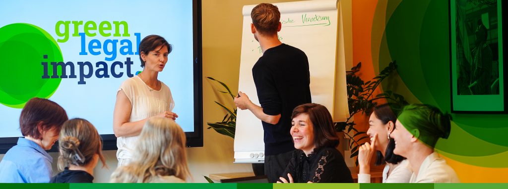 Acht Personen in einer Besprechungssituation: sechs sitzen an einem Tisch, eine Frau steht vor einem Bildschirm und erklärt etwas, neben ihr steht ein Mann und schreibt etwas auf ein Flipchart.
