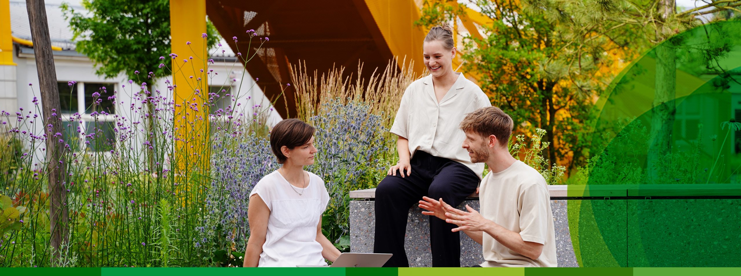 Zwei Frauen und ein Mann sitzen auf Bänken in einem Garten und diskutieren etwas.