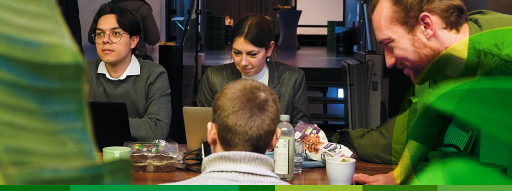 Vier junge Leute arbeiten gemeinsam an einem Tisch, im Vordergrund sind grüne Zimmerpflanzen zu sehen.