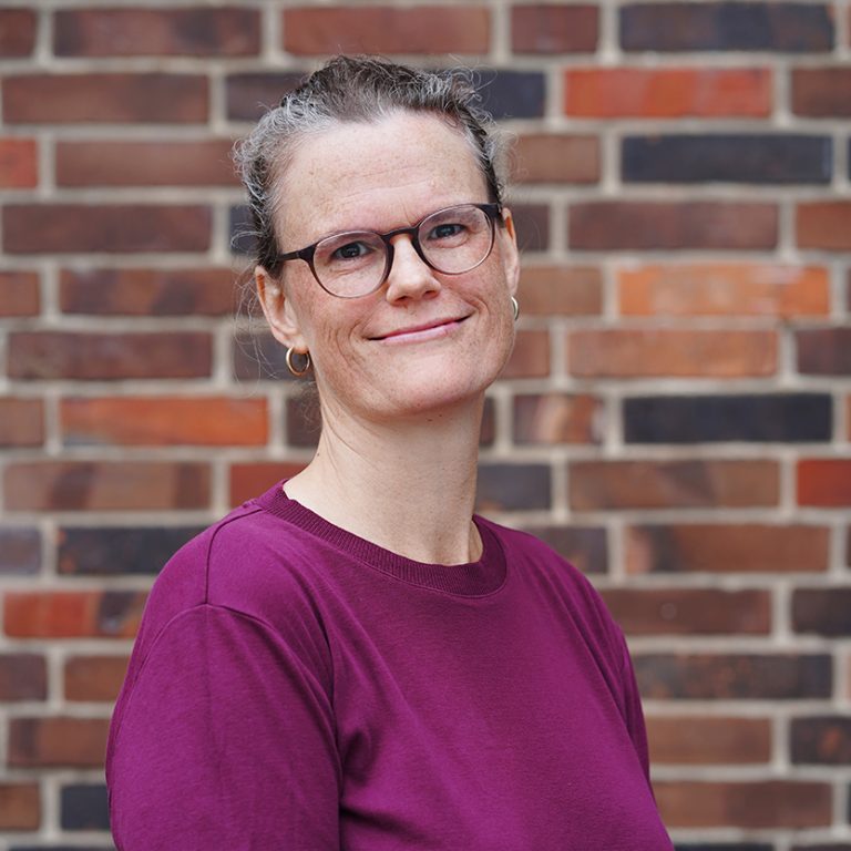 Porträtaufnahme von Johanna Kusch: Eine lächelnde grauhaarige Frau mit Brille, die ein violettes Oberteil trägt.