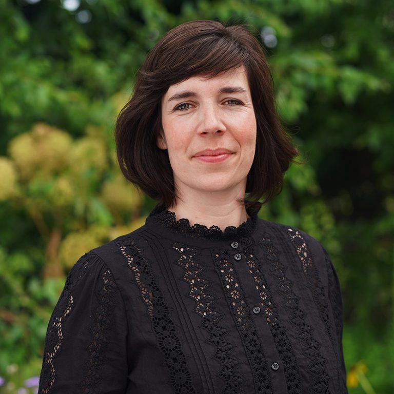 Porträtaufnahme von Judith Hartmann: Eine dunkelhaarige Frau mit dunklen Augen, die eine hochgeschlossene schwarze Bluse trägt.