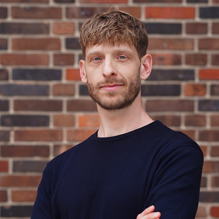 Porträtfoto von Philipp Schönberger: Ein blonder junger Mann mit blauen Augen, der die Arme über seinem dunkelblauen Pullover verschränkt hat.