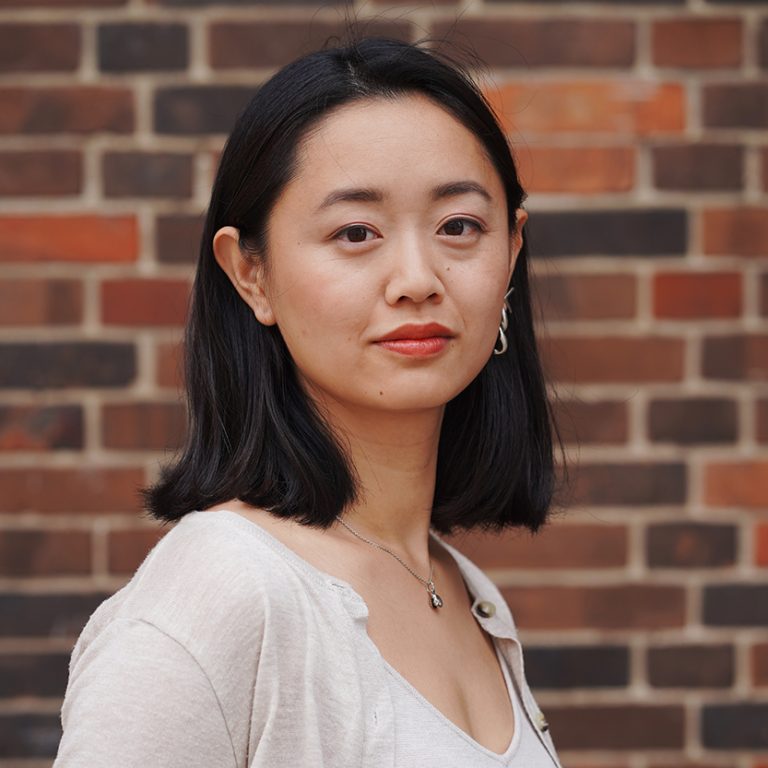 Porträtfoto von Valentine Zheng: eine junge Frau mit schulterlangem schwarzen Haar vor einer Backsteinwand. Sie trägt ein beiges Oberteil, eine Silberkette und einen großen silbernen Ohrring im linken Ohr.