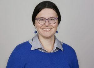 Porträt einer lachenden dunkelhaarigen Frau mit Brille, die einen azurblauen Pullover und farblich passende auffallende Ohrringe aus Glas trägt.