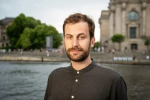 Porträt eines jungen Mannes mit braunem Haar und Bart. Im Hintergrund ist das Reichtagsufer der Spree in Berlin zu sehen.