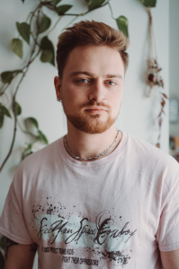 Porträt einer nichtbinären Person mit rotblondem kurzen Haar und Bart und grauen Augen.