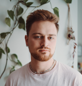 Porträt einer nichtbinären Person mit rotblondem kurzen Haar und Bart und grauen Augen.