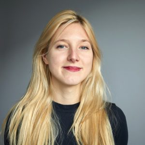 Porträt einer jungen Frau mit langem hellblondem Haar und grauen Augen, die verschmitzt lächelt.