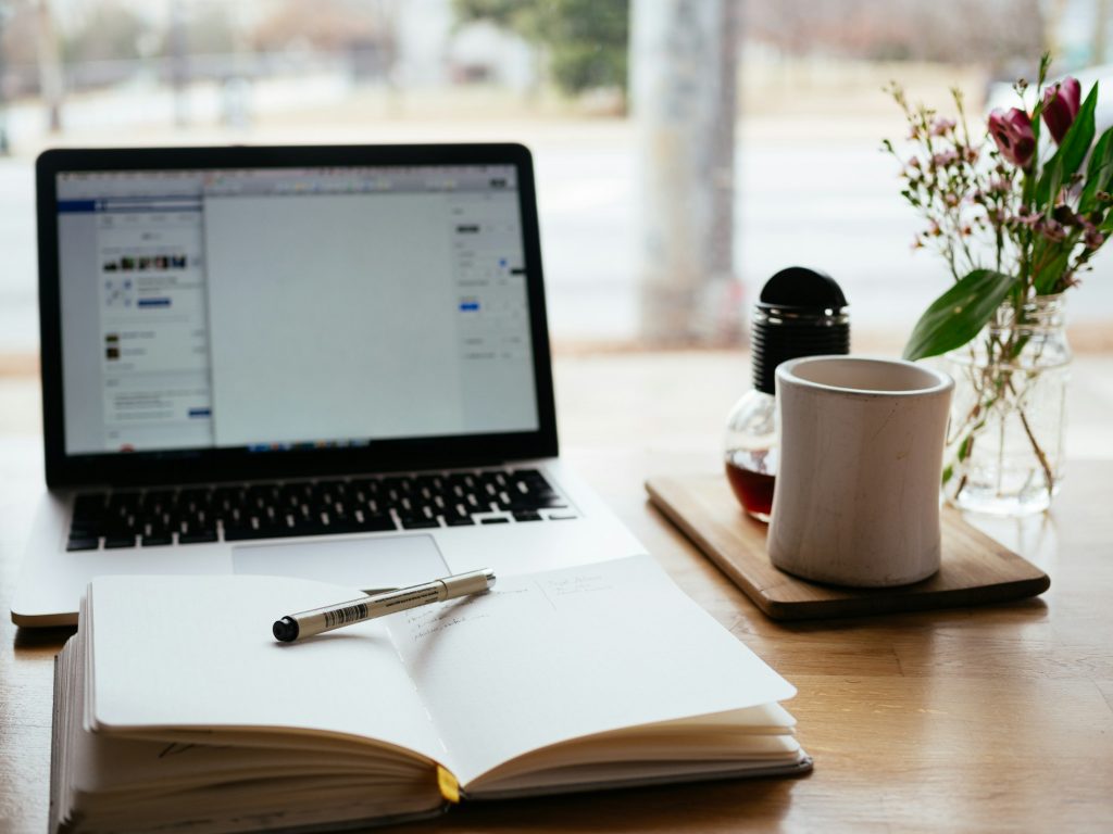 Notizbuch und Stift liegen auf einem Schreibtisch vor einem aufgeklappten Laptop. Rechts daneben steht eine weiße Teetasse auf einem Holzuntersetzer.