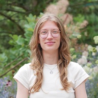 Portrait photo of Kora Stehr: A young woman with blonde hair and glasses against the backdrop of a garden landscape.